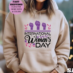 May include: Sand-colored hoodie with a front graphic celebrating International Women's Day. The design features three raised fists in purple, the text "INTERNATIONAL Women's DAY", and floral accents. The hoodie has a drawstring hood and a front pocket.