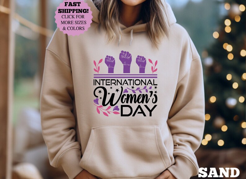 May include: Sand-colored hoodie with a front graphic celebrating International Women's Day. The design features three raised fists in purple, the text "INTERNATIONAL Women's DAY", and floral accents. The hoodie has a drawstring hood and a front pocket.