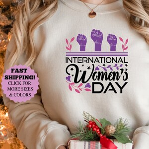 May include: Sand-colored sweatshirt with the text "INTERNATIONAL Women's DAY" in black and purple. The design includes three raised fists and floral accents. A gift box with a festive arrangement is held in the person's hands.