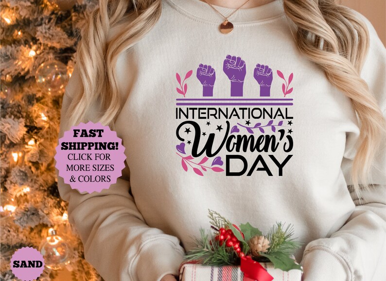 May include: Sand-colored sweatshirt with the text "INTERNATIONAL Women's DAY" in black and purple. The design includes three raised fists and floral accents. A gift box with a festive arrangement is held in the person's hands.