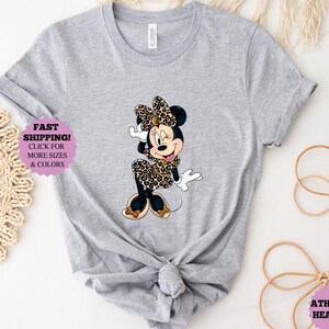 May include: Athletic heather gray t-shirt featuring a Minnie Mouse graphic in a leopard print dress and bow. The shirt has a knotted front. Text on the shirt reads "FAST SHIPPING! CLICK FOR MORE SIZES & COLORS" and "ATHLETIC HEATHER".
