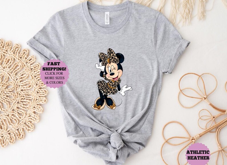 May include: Athletic heather gray t-shirt featuring a Minnie Mouse graphic in a leopard print dress and bow. The shirt has a knotted front. Text on the shirt reads "FAST SHIPPING! CLICK FOR MORE SIZES & COLORS" and "ATHLETIC HEATHER".