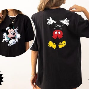 Peut inclure: T-shirt noir avec un motif Mickey Mouse &agrave; l'avant et &agrave; l'arri&egrave;re. Le motif avant montre Mickey per&ccedil;ant un trou d&eacute;chir&eacute;. Le motif arri&egrave;re affiche le visage, les mains et les pieds de Mickey. Le t-shirt porte les mots "Comfort Color Brand 1717" et "Black".