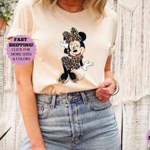 May include: A sand-colored t-shirt featuring a Minnie Mouse graphic in a leopard print dress and bow. The shirt is paired with blue jeans. A purple sticker with the text "FAST SHIPPING! CLICK FOR MORE SIZES & COLORS" is visible.