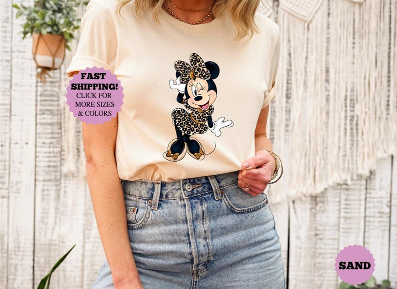 May include: A sand-colored t-shirt featuring a Minnie Mouse graphic in a leopard print dress and bow. The shirt is paired with blue jeans. A purple sticker with the text "FAST SHIPPING! CLICK FOR MORE SIZES & COLORS" is visible.