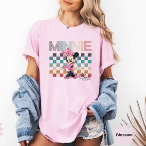 May include: Pink t-shirt featuring a graphic of Minnie Mouse with the word "MINNIE" above her. The design includes a checkered pattern in various colors. The shirt is a Comfort Color brand.