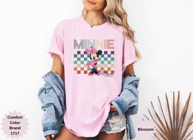 May include: Pink t-shirt featuring a graphic of Minnie Mouse with the word "MINNIE" above her. The design includes a checkered pattern in various colors. The shirt is a Comfort Color brand.