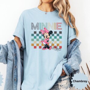 May include: Light blue t-shirt featuring a Minnie Mouse graphic. The design includes the word "MINNIE" in a retro font above a checkered pattern and a Minnie Mouse illustration in a pink dress. The shirt is a Comfort Color Brand.