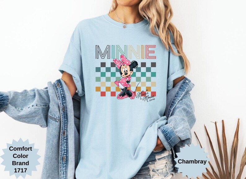 May include: Light blue t-shirt featuring a Minnie Mouse graphic. The design includes the word "MINNIE" in a retro font above a checkered pattern and a Minnie Mouse illustration in a pink dress. The shirt is a Comfort Color Brand.