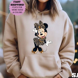 May include: Sand-colored hoodie featuring a graphic of Minnie Mouse in a leopard print outfit. The image includes the text "FAST SHIPPING! CLICK FOR MORE SIZES & COLORS" in a pink circle. The word "SAND" is printed at the bottom.