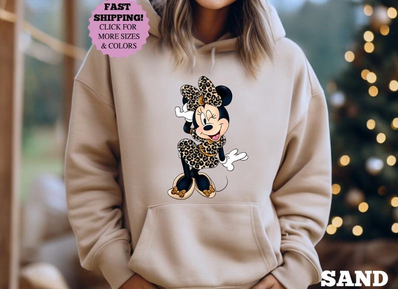 May include: Sand-colored hoodie featuring a graphic of Minnie Mouse in a leopard print outfit. The image includes the text "FAST SHIPPING! CLICK FOR MORE SIZES & COLORS" in a pink circle. The word "SAND" is printed at the bottom.