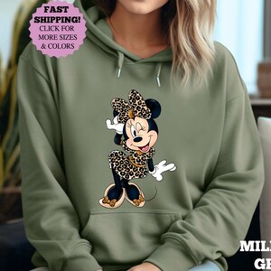 May include: Olive green hooded sweatshirt featuring a Minnie Mouse graphic in a leopard print dress and bow. The sweatshirt has a front pocket and drawstring hood. The text "MILITARY GREEN" is visible on the bottom right.