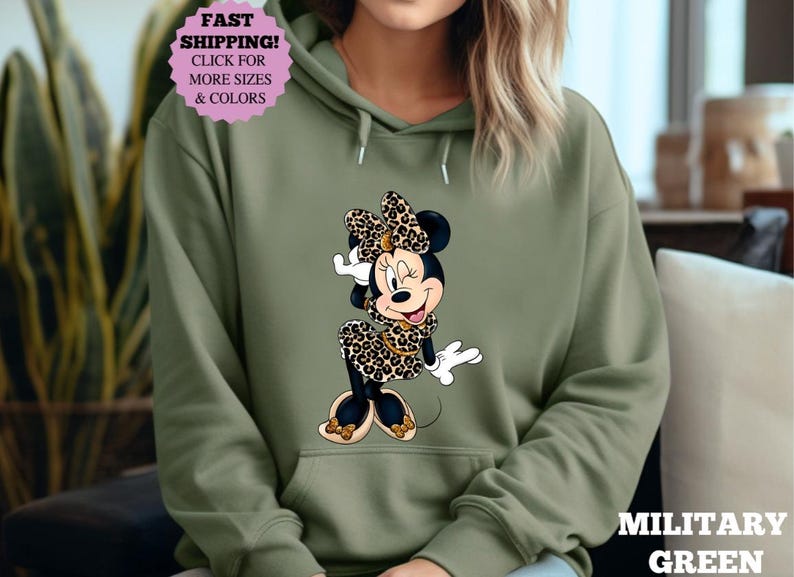 May include: Olive green hooded sweatshirt featuring a Minnie Mouse graphic in a leopard print dress and bow. The sweatshirt has a front pocket and drawstring hood. The text "MILITARY GREEN" is visible on the bottom right.
