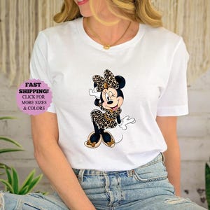 May include: White t-shirt featuring a Minnie Mouse graphic in a leopard print design. The cartoon character is wearing a matching bow, dress, and shoes. The shirt also has a purple sticker that says "FAST SHIPPING! CLICK FOR MORE SIZES & COLORS."