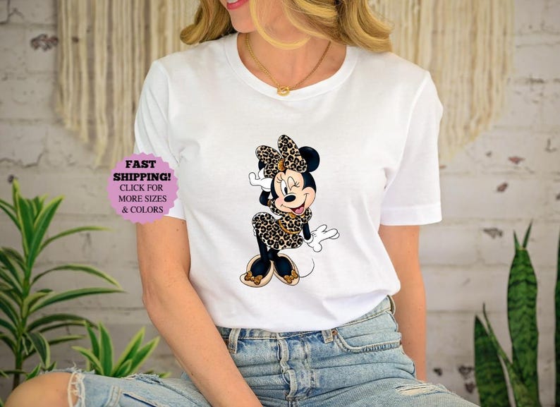 May include: White t-shirt featuring a Minnie Mouse graphic in a leopard print design. The cartoon character is wearing a matching bow, dress, and shoes. The shirt also has a purple sticker that says "FAST SHIPPING! CLICK FOR MORE SIZES & COLORS."