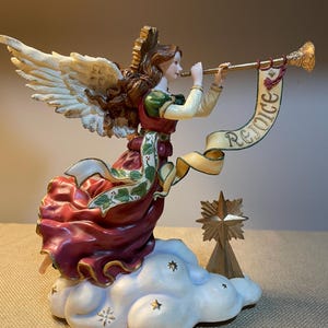 May include: A decorative angel figurine with outstretched wings, playing a golden trumpet. The angel wears a red and green dress and is perched on a cloud. A banner reads "Rejoice". A wooden star is also present.