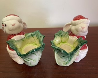 May include: Two white ceramic candle holders shaped like bunnies wearing Santa hats. Each bunny is holding a green and white ceramic leaf-shaped candle holder.