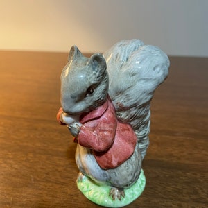 May include: A gray and brown squirrel figurine wearing a red jacket, holding a nut in its paws. The figurine is standing on a green base.