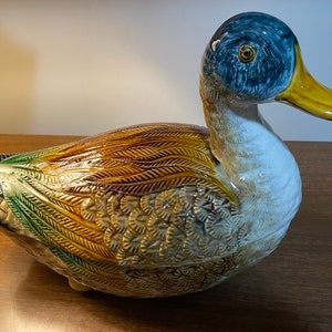 May include: A ceramic duck figurine with brown, green, and blue painted details. The duck has a detailed textured surface and a yellow beak.