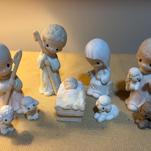 May include: A porcelain nativity set featuring figures of a baby in a manger, two shepherds holding staffs, a seated figure, and four small lambs. The figures have matte white finishes with subtle pastel accents on the clothing and blankets.
