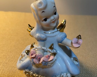 May include: A porcelain figurine of a girl angel with blue wings, a blue dress, and a pink flower hat. She is holding a basket of pink flowers and the base of the figurine says "Friday's Child".