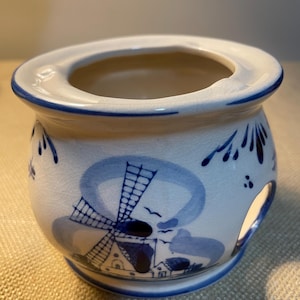 May include: A white ceramic candle holder with blue painted windmill and floral designs. The holder has a round shape with a wide opening and a blue rim. The design is reminiscent of Delftware.