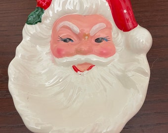 May include: A white ceramic Santa Claus dish with a red hat, white beard, and a green holly sprig.