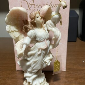May include: A white porcelain figurine of an angel with pink wings, holding a golden trumpet. The angel is standing on a cloud and has a pink ribbon around her waist. The figurine is labeled "Seraphim" and has a gold tag with the letter "R" on it.