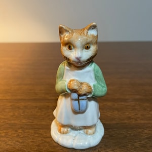 May include: A ceramic figurine of a ginger cat wearing a green jacket and white apron, holding a blue and white gift box.