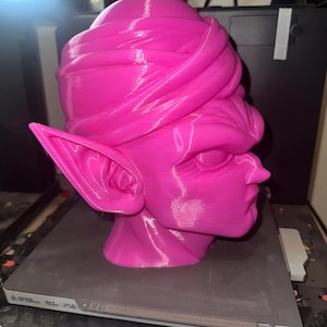May include: A vibrant pink 3D-printed bust of a character with large ears and a turban-like head covering. The bust is on a gray surface, likely a printer bed, with the printer's components visible in the background. The sculpture has a smooth, glossy finish.