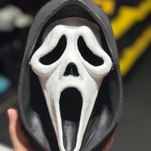 Könnte beinhalten: Eine Scream-Maske mit einem weißen, länglichen Gesicht, großen ovalen Augenöffnungen und offenem Mund. Die Maske wird von einer schwarzen Kapuze umrahmt, was einen starken Kontrast erzeugt. Ein beliebtes Halloween-Kostümzubehör.