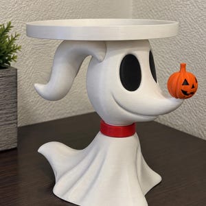 May include: A white ghost-shaped stand featuring a flat circular top, large black oval eyes, and a red collar. A small orange jack-o'-lantern with a carved face is balanced on the tip of the ghost's nose. The figure has a flowing base.