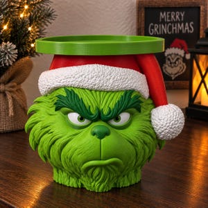 Puede incluir: Una maceta con forma de cabeza de personaje verde texturizada, con el ceño fruncido y expresión de enfado. La figura lleva un gorro de Papá Noel rojo con borde blanco texturizado y un pompón blanco. Un borde circular verde se asienta sobre el gorro.
