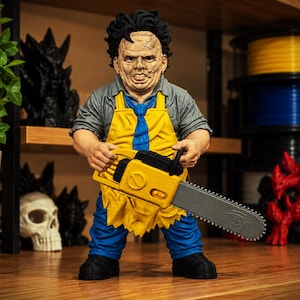 May include: A figurine of a character with a stitched mask, black hair, and a grey shirt, wearing a bright yellow apron and a blue tie. The figure holds a yellow and grey chainsaw with both hands and wears blue pants with black shoes.