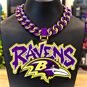Könnte beinhalten: Eine klobige Kettenhalskette mit ineinandergreifenden violetten und gelben Gliedern. Ein großer Anhänger hängt in der Mitte und zeigt das Wort RAVENS in violetten Graffiti-Buchstaben mit gelben Umrandungen über einem stilisierten Rabenkopf-Logo.