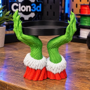 May include: A decorative tray stand featuring two sculpted green furry hands emerging from red and white cuffs, holding a circular black tray. The hands are textured to resemble fur, and the base consists of two red sleeves with white trim.