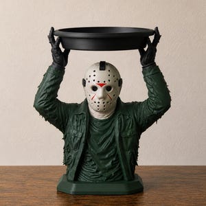Peut inclure: Une figurine décorative représentant un personnage portant un masque de hockey blanc avec des accents rouges et une veste vert foncé usée. Le personnage a les deux bras levés au-dessus de la tête, tenant un plateau noir circulaire.