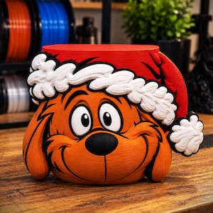 Puede incluir: Una maceta con forma de criatura naranja impresa en 3D, con una nariz negra grande, ojos abiertos y un gorro de Papá Noel rojo con borde y pompón blancos texturizados. El objeto tiene una forma redondeada con líneas de impresión visibles.