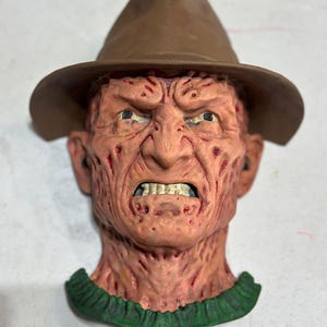 Könnte beinhalten: Detaillierte Maske einer Figur mit braunem Fedora-Hut und grünem Kragen. Die Maske hat ein verwittertes, vernarbtes Gesicht mit wütendem Ausdruck und zeigt Zähne. Der Hautton ist eine Mischung aus Rosa und Rot.