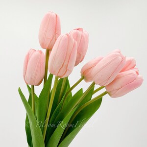 Puede incluir: Un ramo de tulipanes rosas artificiales con tallos y hojas verdes. Los tulipanes se sostienen en una mano sobre un fondo blanco. La imagen incluye el texto "Bloom Room Floral".