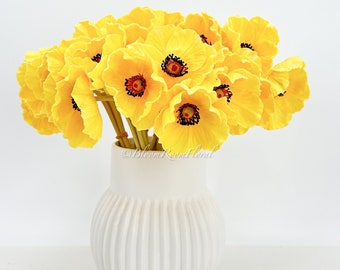 Puede incluir: Un ramo de flores de amapola de seda de color amarillo brillante en un jarrón de cerámica blanco acanalado. Las flores tienen centros negros con acentos rojos.