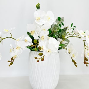 May include: White orchid flower arrangement in a white textured vase. The orchids are arranged in a cascading style with green leaves.