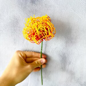 K&ouml;nnte beinhalten: Eine einzelne k&uuml;nstliche Blume mit einem gelben und orangenen Bl&uuml;tenkopf. Der Bl&uuml;tenkopf besteht aus d&uuml;nnen, gekr&auml;uselten Dr&auml;hten. Der Bl&uuml;tenstiel ist gr&uuml;n.