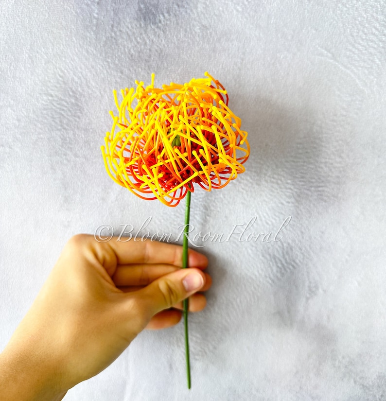 K&ouml;nnte beinhalten: Eine einzelne k&uuml;nstliche Blume mit einem gelben und orangenen Bl&uuml;tenkopf. Der Bl&uuml;tenkopf besteht aus d&uuml;nnen, gekr&auml;uselten Dr&auml;hten. Der Bl&uuml;tenstiel ist gr&uuml;n.