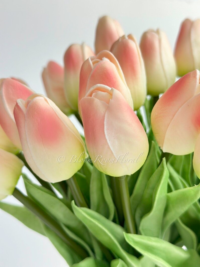 May include: A bouquet of artificial tulips with pink and white petals and green leaves. The tulips are arranged in a vase, and the stems are visible.