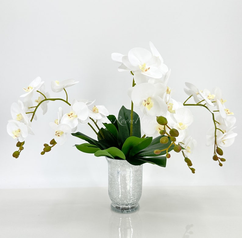 May include: White orchid flower arrangement in a silver vase. The orchids have white petals and yellow centers. The vase is a silver color with a textured surface.