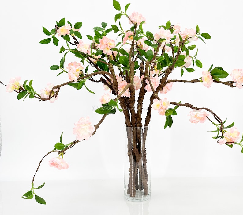 May include: A clear glass vase holds a floral arrangement of faux cherry blossom branches. The branches have light pink flowers, green leaves, and brown stems. The arrangement is set against a white background.