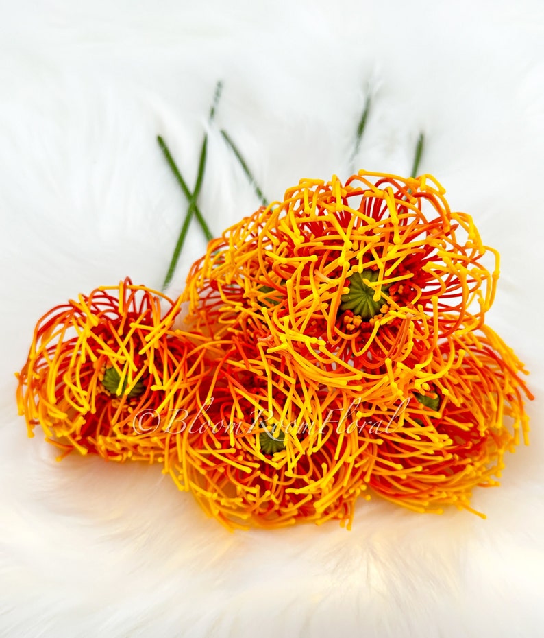 K&ouml;nnte beinhalten: Ein Strau&szlig; aus orangefarbenen und gelben Kunstblumen mit drahtartiger Textur. Die Blumen sind in einer Gruppe angeordnet und haben ein zartes, federleichtes Aussehen.