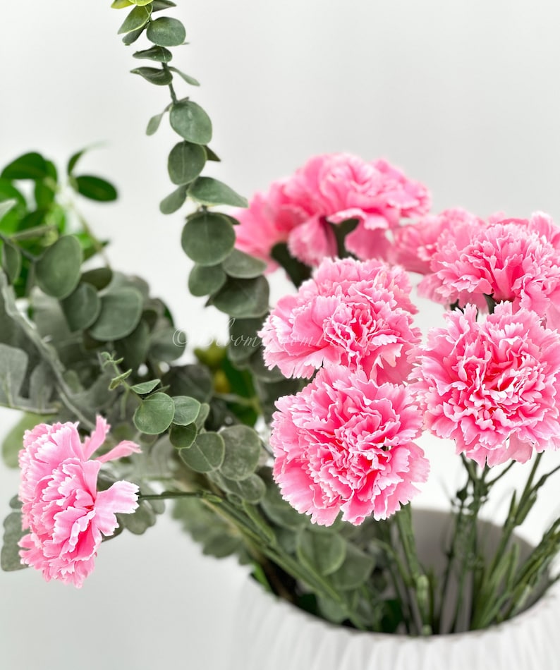 May include: A bouquet of pink carnations with green eucalyptus leaves in a white vase.