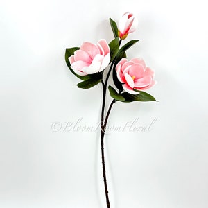 May include: A single stem of artificial pink magnolia flowers with green leaves. The stem is brown and has a natural look.